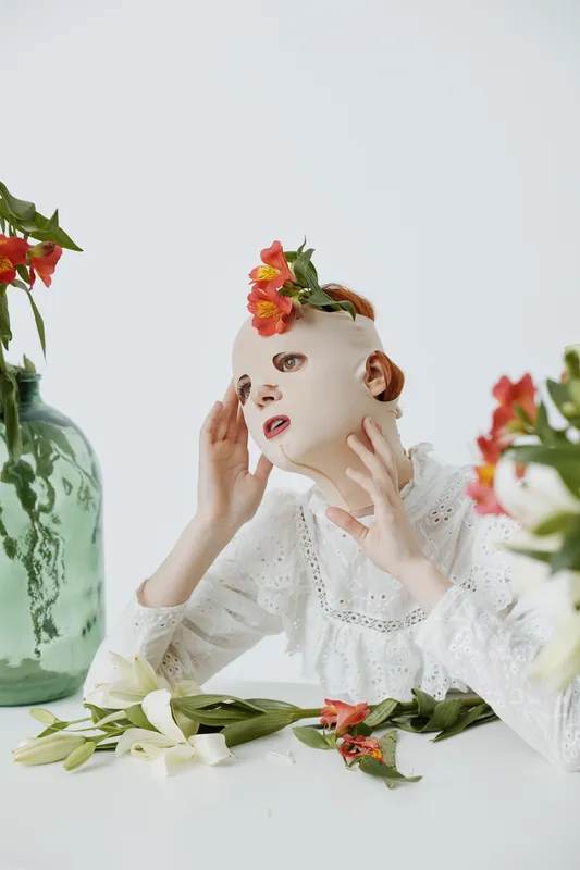 Young model with a mask adorned with flowers, expressing emotion in a minimalist setting, perfect for fashion and beauty concepts
