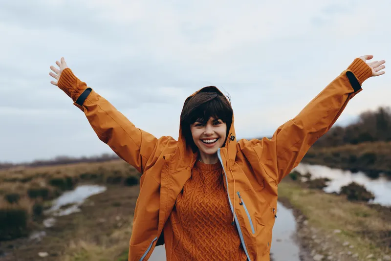 Woman outdoors smiling on a hiking trail, arms raised in joy, wearing orange jacket and knit sweater, lifestyle in nature, autumn landscape under soft sky light, cloudy sky and hills
