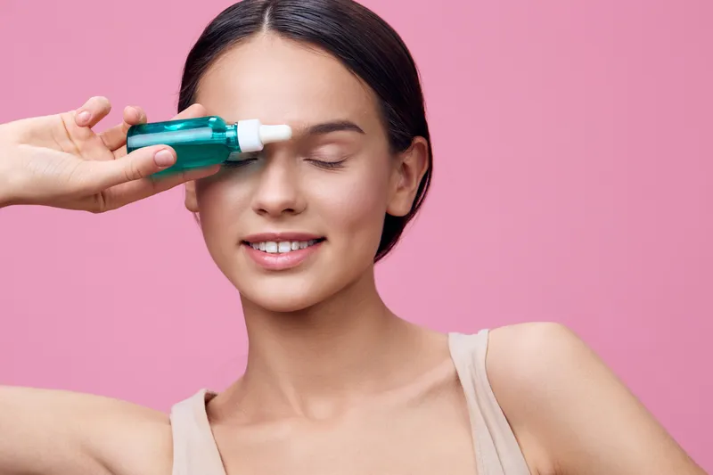 Young woman applying skincare serum with a joyful expression against a pink background for healthy skin and beauty concepts