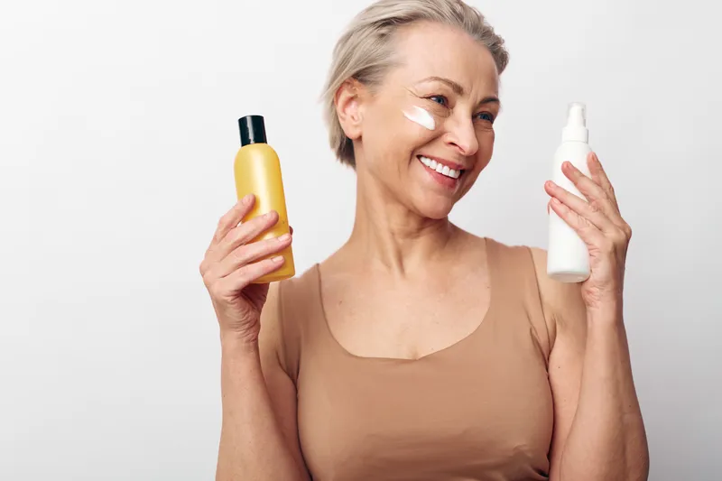 Smiling middle aged woman with short hair holding two skincare bottles and applying cream on cheek, isolated on white background. Concept of beauty routine, self care, and mature skin wellness.