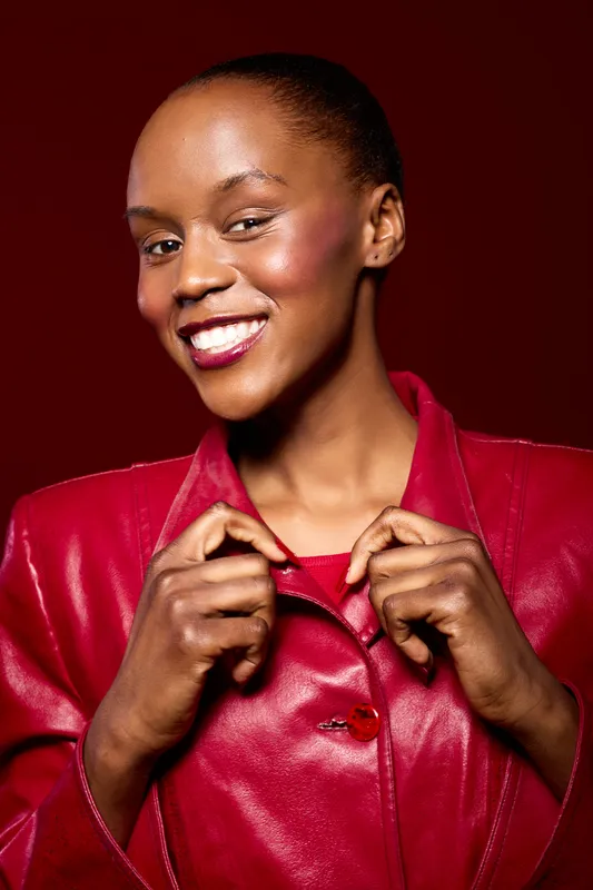 Smiling African woman with dark skin wearing a red leather jacket poses confidently against a red background. Concept of fashion, beauty, and empowerment with vibrant colors.
