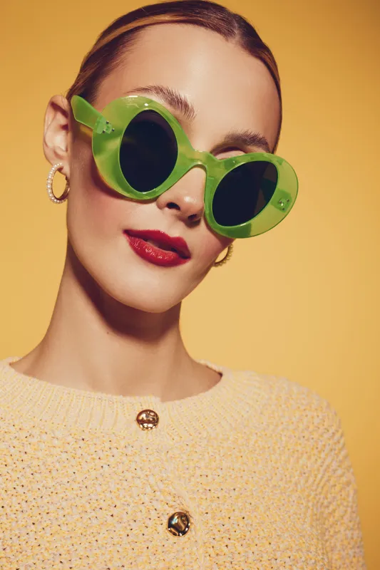 Stylish woman with green round sunglasses and red lipstick poses against yellow background. She wears a textured sweater with gold buttons and hoop earrings. Fashion and beauty concept.