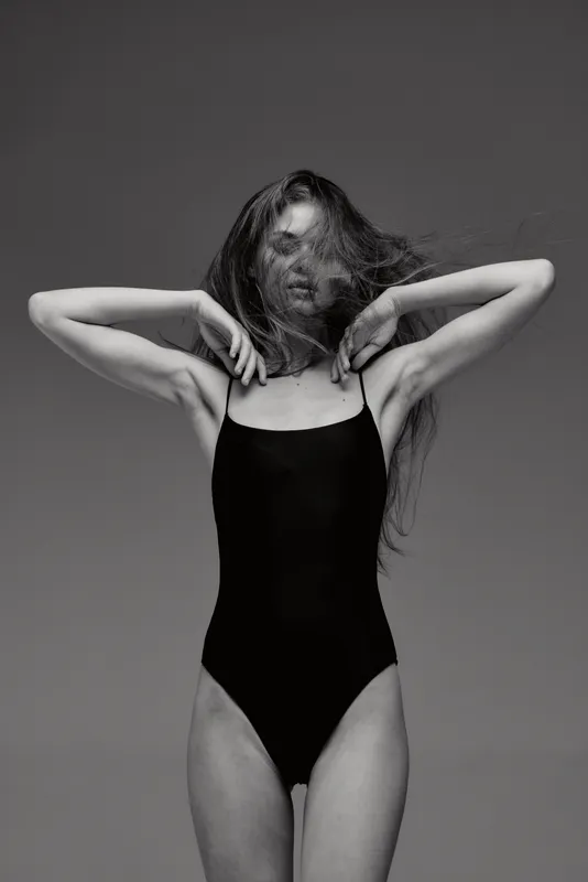 Black and white photo of a woman in a black bodysuit posing with arms raised and hair flowing, artistic and expressive portrait.