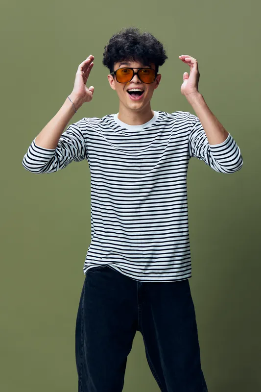 young man wearing striped shirt and orange sunglasses joyfully expressing surprise with raised hands on olive green background, casual and cheerful mood
