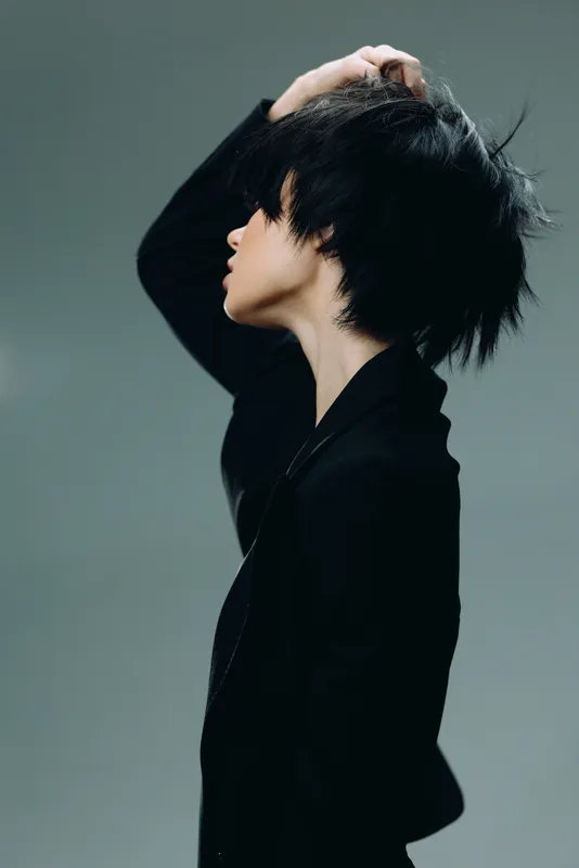 Profile of stylish woman with short black hair wearing black coat, posing thoughtfully against dark gray background.