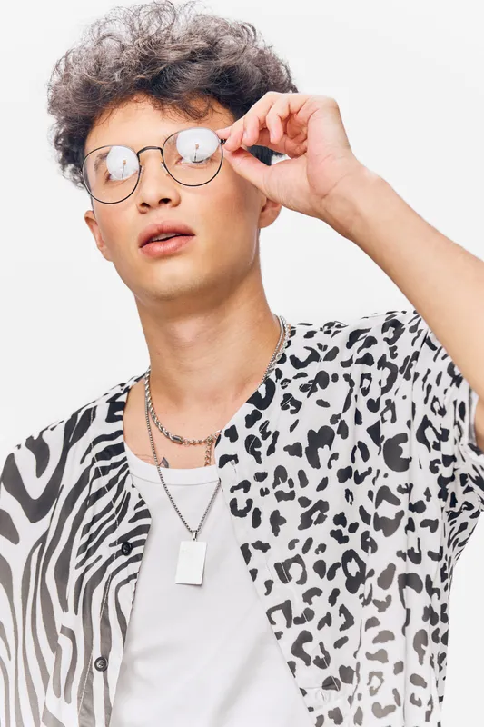 young man in stylish animal print shirt adjusting round glasses with confident expression against white background in bright studio setting
