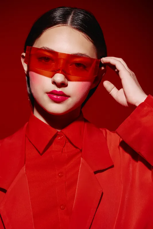 Stylish young woman wearing red translucent sunglasses and a red suit poses confidently against a vivid red background. Concept of bold beauty and fashion creativity.