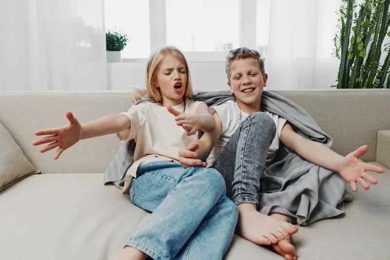 Two playful children sitting on sofa singing or acting with expressive faces and open arms in bright living room interior.