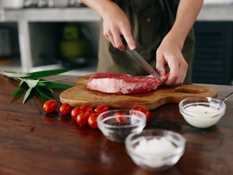 Женщина с ножом в руке нарезает свежее стейковое мясо для запекания на кухне