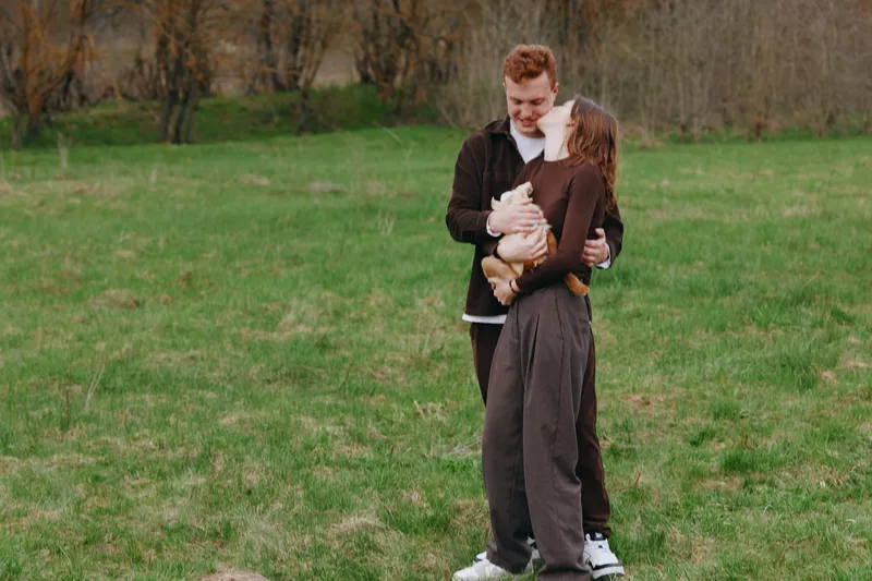 A joyful couple outdoors in a grassy field cradle a small goat kid, sharing a warm moment that celebrates family bonds, countryside life, and gentle animal companionship.