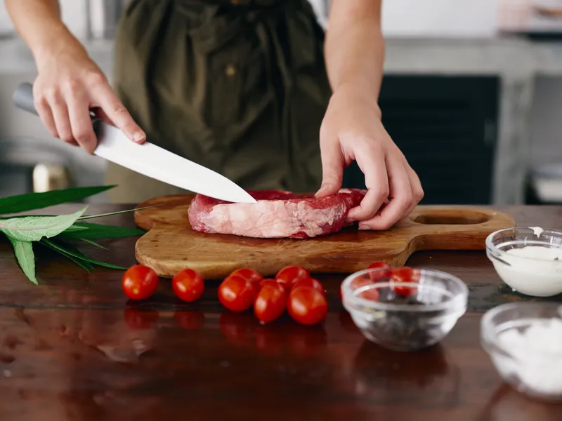 Женщина с ножом в руке режет свежее сырое мясо для жарки на кухне