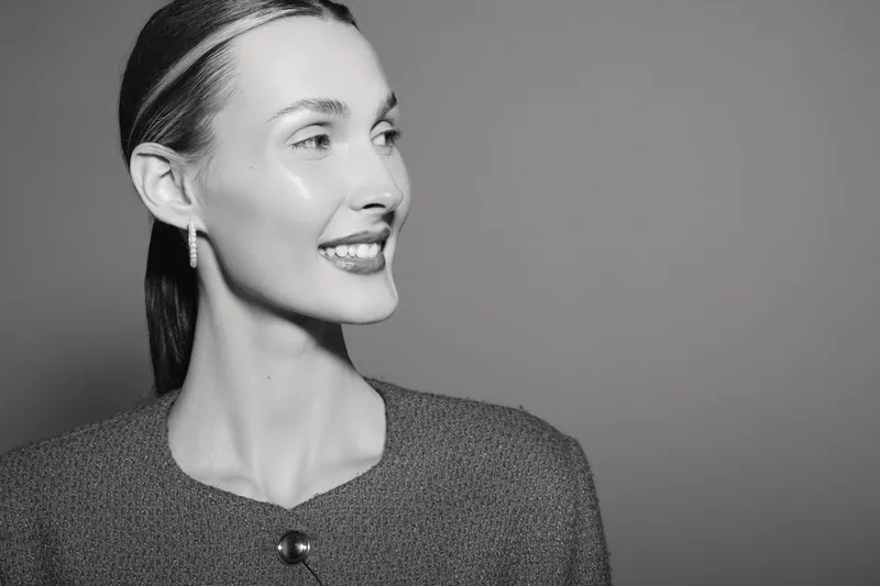 Smiling Caucasian woman with pale skin and slicked back hair wearing textured top and earrings looks sideways on grey background. Concept of beauty, fashion, and elegance.