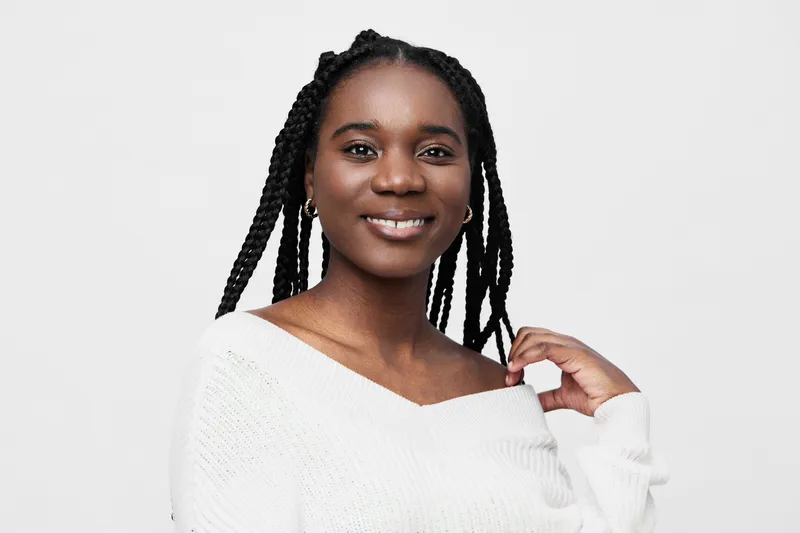 Smiling young woman with braided hair wearing a cozy white sweater, posing against a neutral background, conveying warmth and positivity in a minimalist style