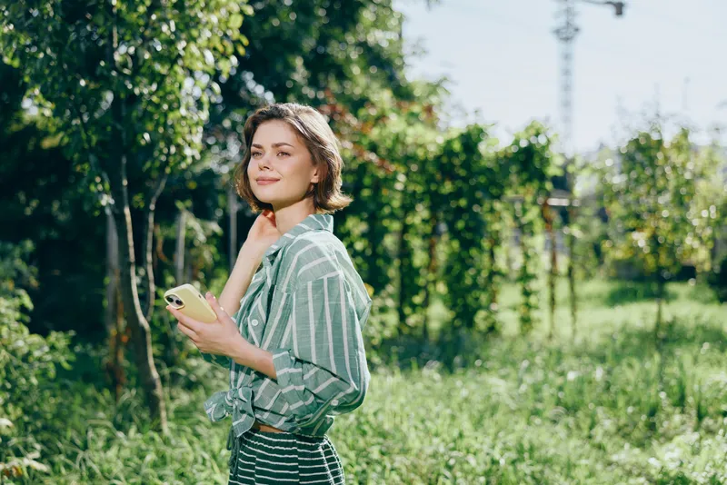 A young woman stands in a sunny garden, glancing upward with a thoughtful expression while holding a smartphone.