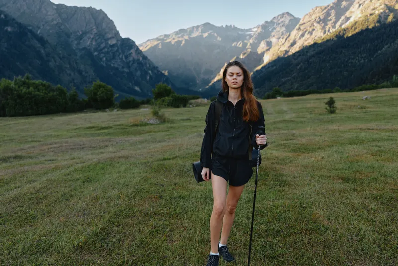 A young woman hikes through a grassy valley framed by towering mountains. She wears outdoor gear and a trekking pole, gazing ahead with calm focus amid vast natural scenery.