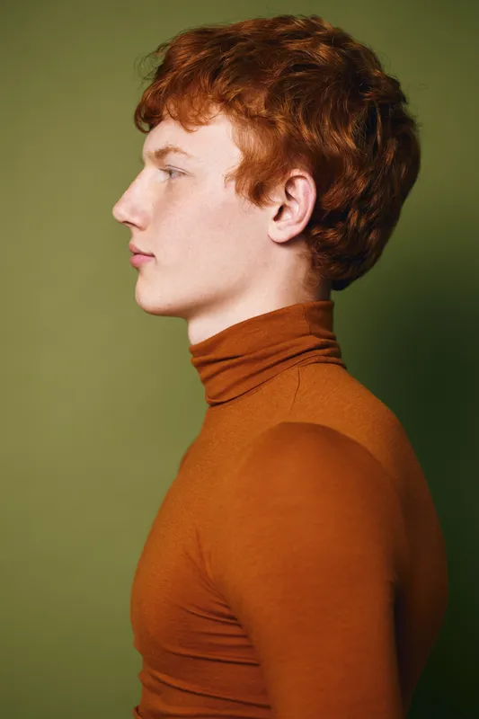 Portrait of young man with red hair wearing brown turtleneck sweater looking sideways against plain green background. Profile view, casual clothing and soft lighting in studio shot.