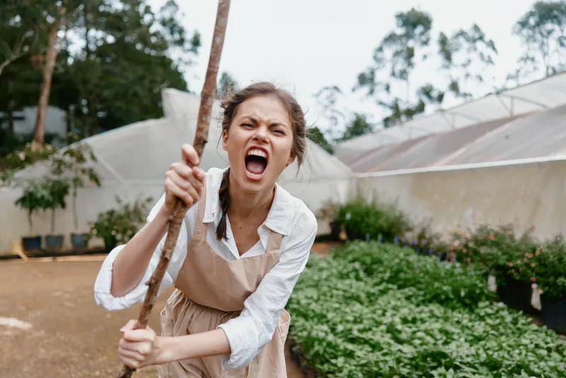 Young woman with brown hair expressing anger while holding a stick in a garden setting, wearing an apron and white shirt, surrounded by green plants
