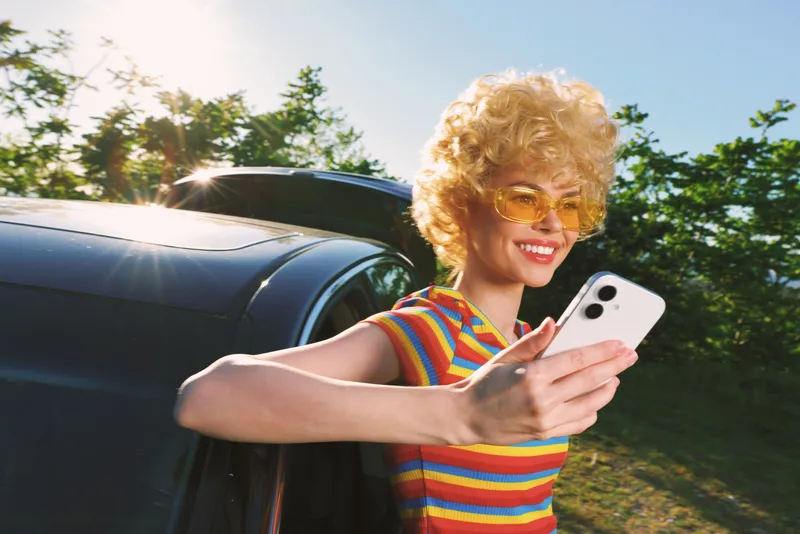 Woman lifestyle scene with retro color palette, posing by car window, taking a selfie, wearing striped shirt and yellow sunglasses, bright sunny day, candid cheerful moment