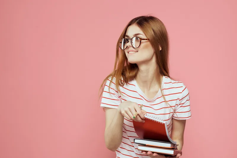 Молодая студентка с книгами на розовом фоне, в очках на лице, кадрированный вид, учебное заведение