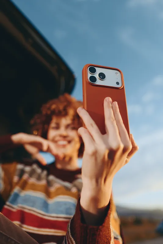 Woman takes a selfie with a smartphone outdoors, lifestyle moment with young smiling expression, hands hold the phone toward the camera, casual sweater under sunlight, background landscape