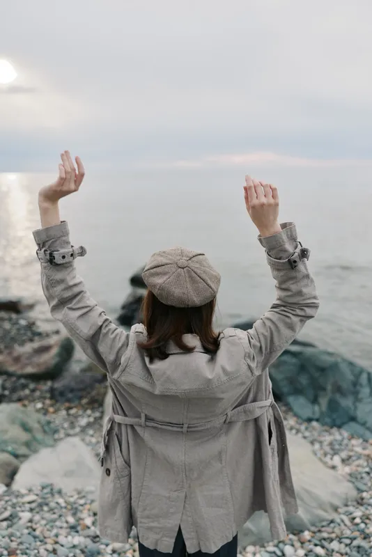 Woman in a coat and hat raises her arms while standing on a rocky beach. Overcast sky and calm sea create a serene and contemplative atmosphere in nature.