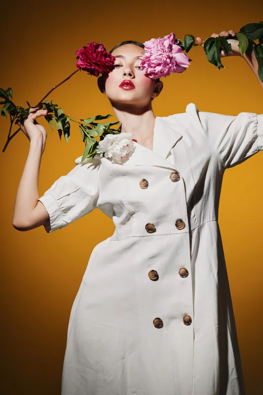 Young woman in white dress holds vibrant pink and red flowers near her face. Bright orange background enhances the fashion beauty concept with creative posing and stylish mood.