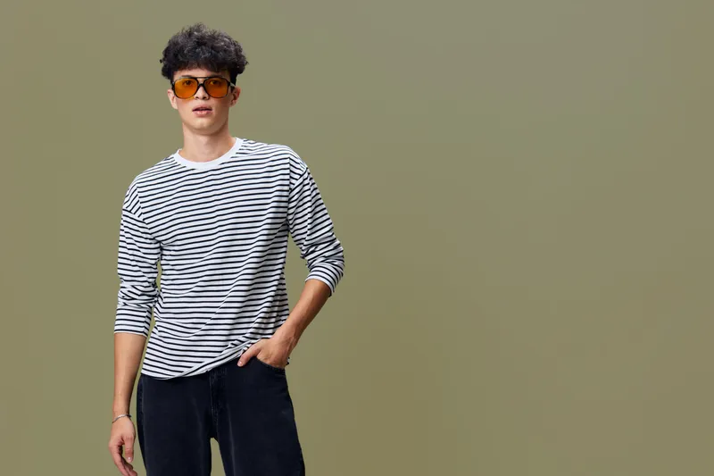 young man wearing striped shirt and sunglasses stands confidently against plain olive background, casual style with cool summer vibes and relaxed posture