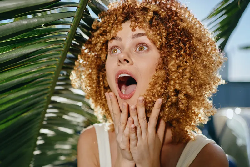 Surprised woman with curly hair expressing shock and amazement near green palm leaves under bright sunlight, showing natural beauty and vibrant emotions.