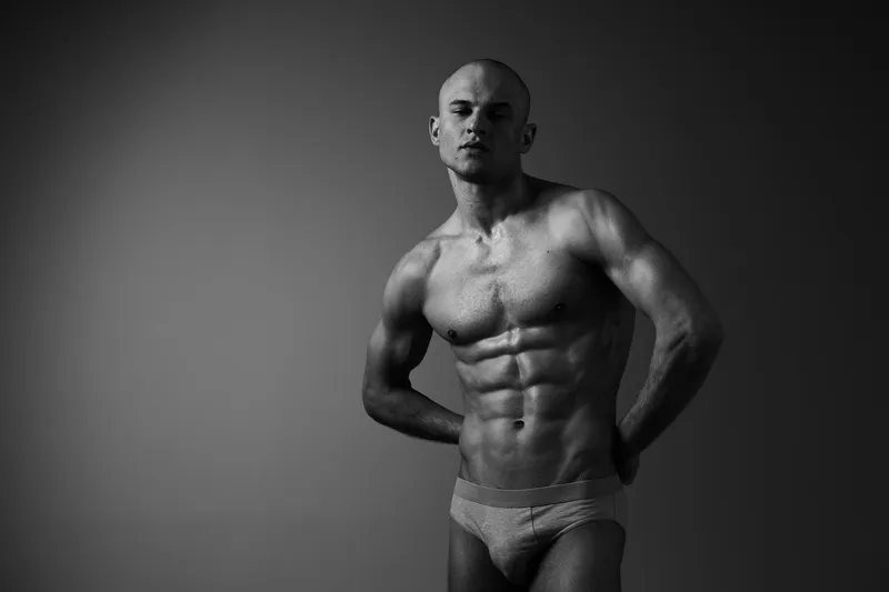 Muscular man posing in black and white, showcasing defined abs and strong physique, exuding confidence and strength in an artistic composition.