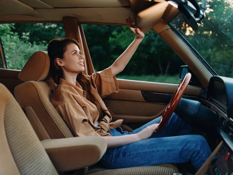 Young woman smiling in a vintage car, enjoying the ride with a relaxed posture and casual outfit, surrounded by greenery and warm sunlight, conveying freedom and adventure.