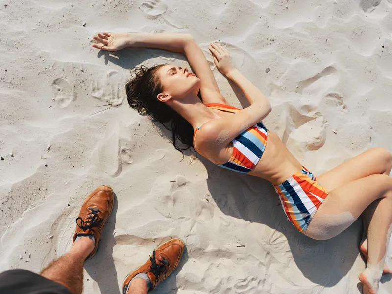 Sunlit beach scene featuring a woman in a colorful striped bikini resting on soft white sand, arms raised and relaxed, toes sink into warm shore.