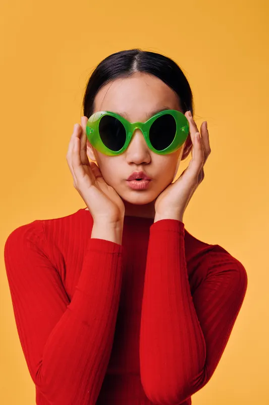 Young woman wearing oversized green sunglasses and red sweater posing on yellow background. Stylish female portrait with playful expression, trendy fashion accessory and colorful studio light.