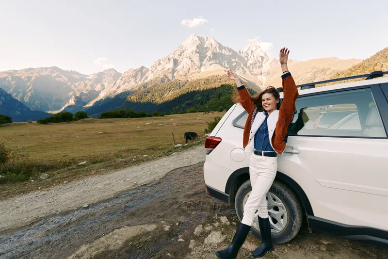 Woman by car in mountains, travel and nature road in countryside with raised arms, smiling and enjoying adventure and scenic view near parked SUV on gravel path.