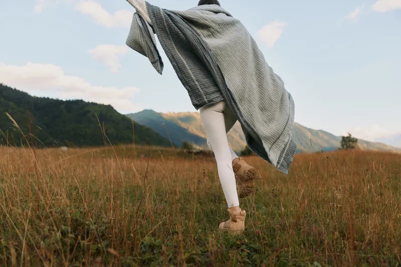 Young woman leaps across a sunlit meadow with a flowing grey shawl, capturing a moment of freedom and carefree outdoor living.