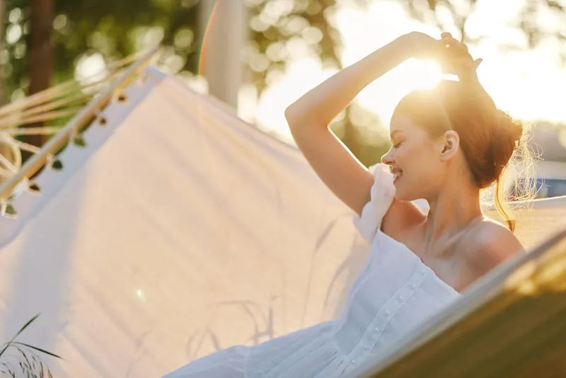 Young woman relaxing in a hammock at sunset, wearing a white dress, showcasing joy and serenity in a tranquil outdoor setting