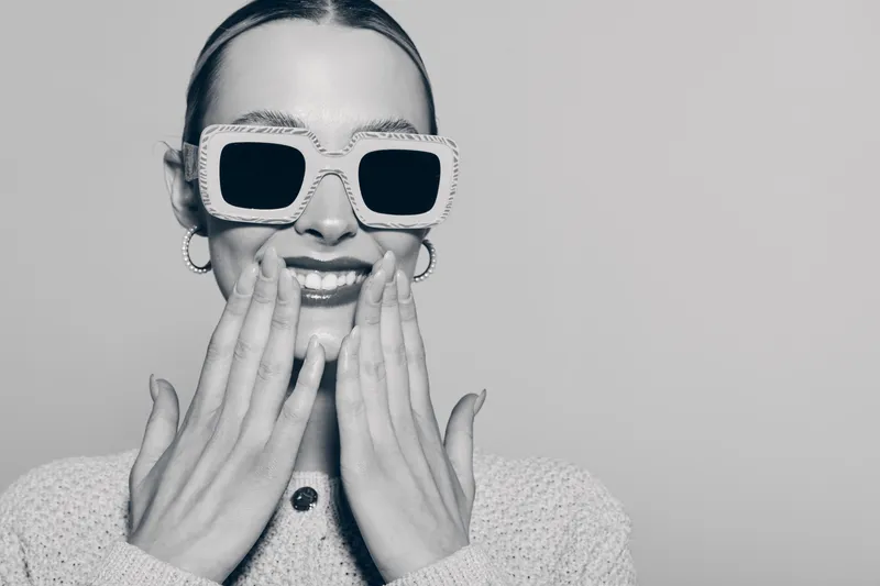 Smiling young white woman with fair skin wears large stylish sunglasses and knitted sweater, covering mouth with hands. Studio portrait with trendy fashion and cheerful mood concept.