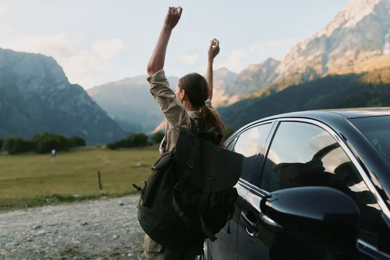 A woman traveler stands beside a car in a rugged mountain valley, backpack on her back, arms raised in triumph, embracing outdoor adventure and freedom.