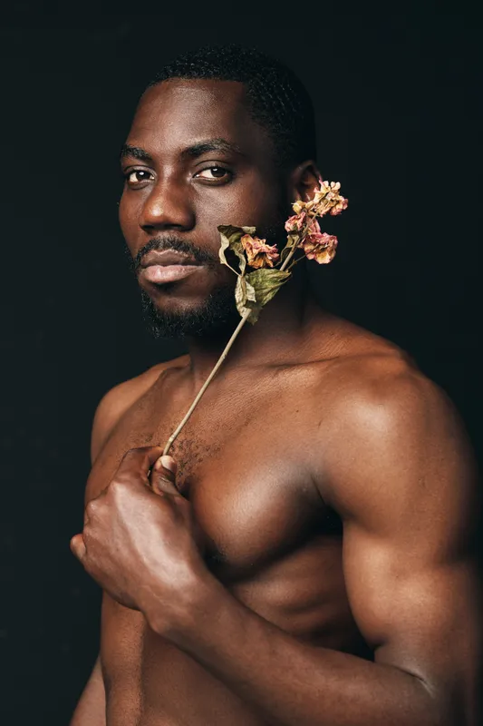 African man with muscular body holding dried flower near face in creative conceptual visual style. Portrait of shirtless black male with serious expression on dark background for artistic photo