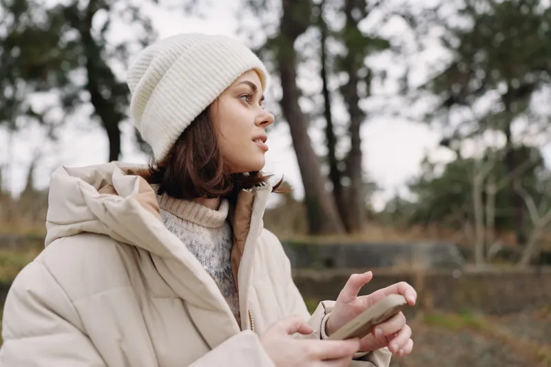 Woman in a beige winter coat and white beanie outdoors, looking to the side while using a phone, in a park with soft natural light.