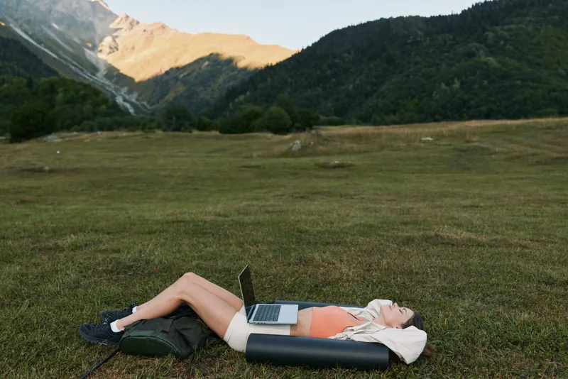 A person lies on a grassy meadow with a laptop resting on their lap in a tranquil outdoor setting. The distant mountains rise, creating a serene work life balance.