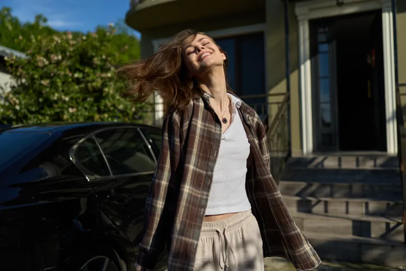 Young woman in a plaid jacket stands beside a car in sunlit street, smiling as her hair blows, creating a casual street fashion moment