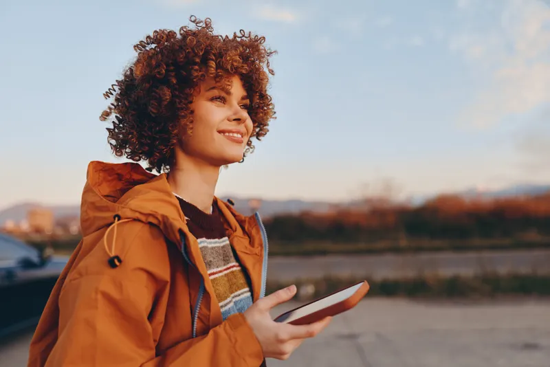 Lifestyle portrait outdoors shows a woman with curly hair wearing an orange jacket, smiling and using a smartphone, warm sunset light, communication and modern daily life