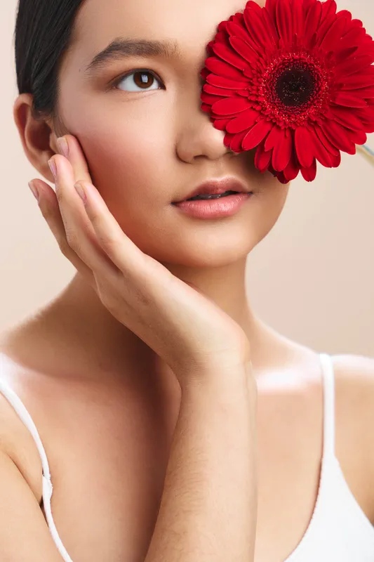 asian woman with a red flower in her hair gazes softly at the camera, minimal makeup, delicate pose, serene mood, beauty portrait for lifestyle and wellness use in marketing contexts.