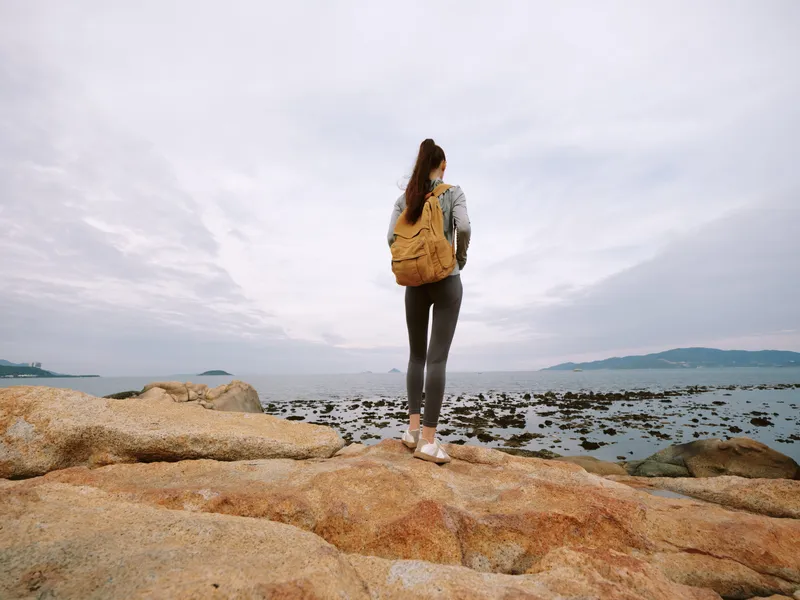 Young woman with a backpack standing on rocky shore, gazing at the sea with serene expression, perfect for travel or adventure concepts