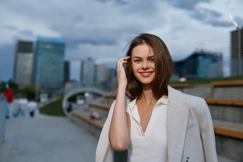 Confident woman in business attire smiling outdoors in urban setting with modern skyscrapers under cloudy sky, casual gray blazer and white blouse