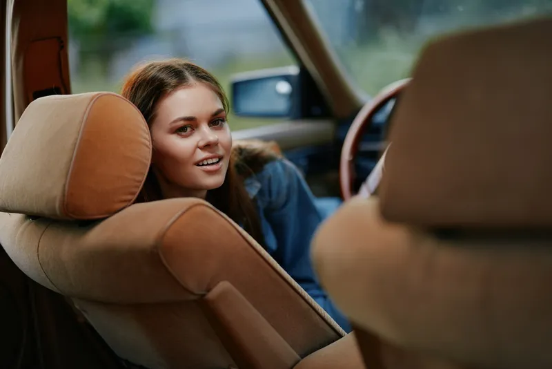 Smiling young woman in a casual outfit sitting in a vintage car, showcasing happiness and comfort in a natural setting.
