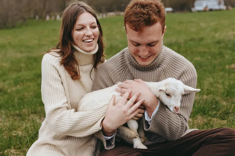 Couple in cozy sweaters cuddle a gentle goat kid in a grassy field, sharing warmth and smiles, a candid countryside moment of affection and relaxed lifestyle.