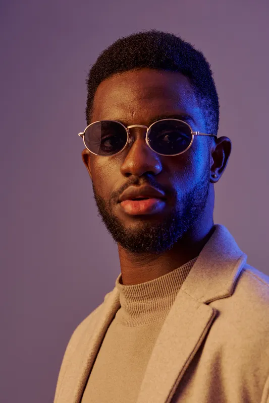 Stylish young Black man wearing sunglasses and a beige blazer against a purple backdrop, exuding confidence and elegance in a modern fashion context