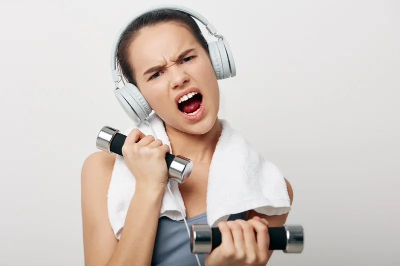 Woman in a fitness scene, lifting dumbbells with headphones on and a towel around her neck, showcasing energy, focus, and gym-ready determination.