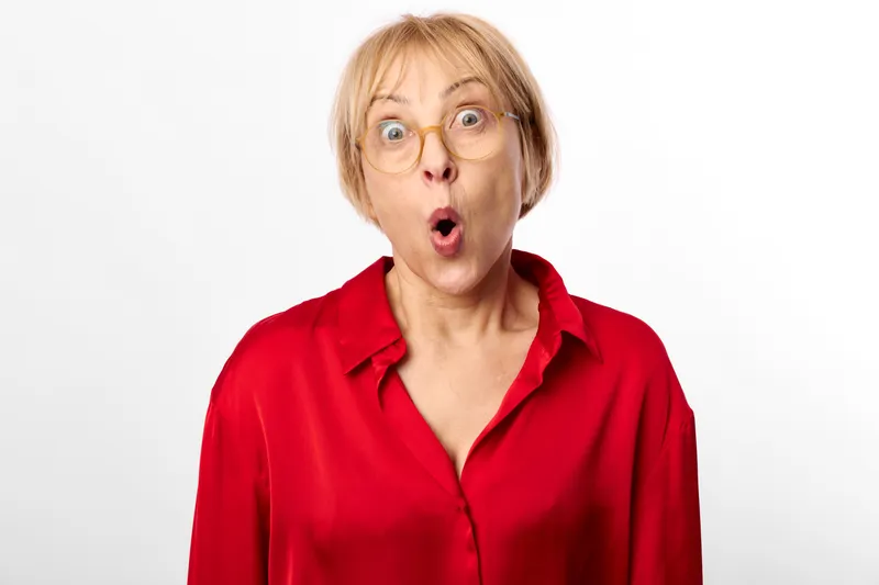 Woman surprised shocked expression glasses open mouth red shirt studio portrait against white background, older adult female with astonished eyes and tense gesture, isolated with copy space